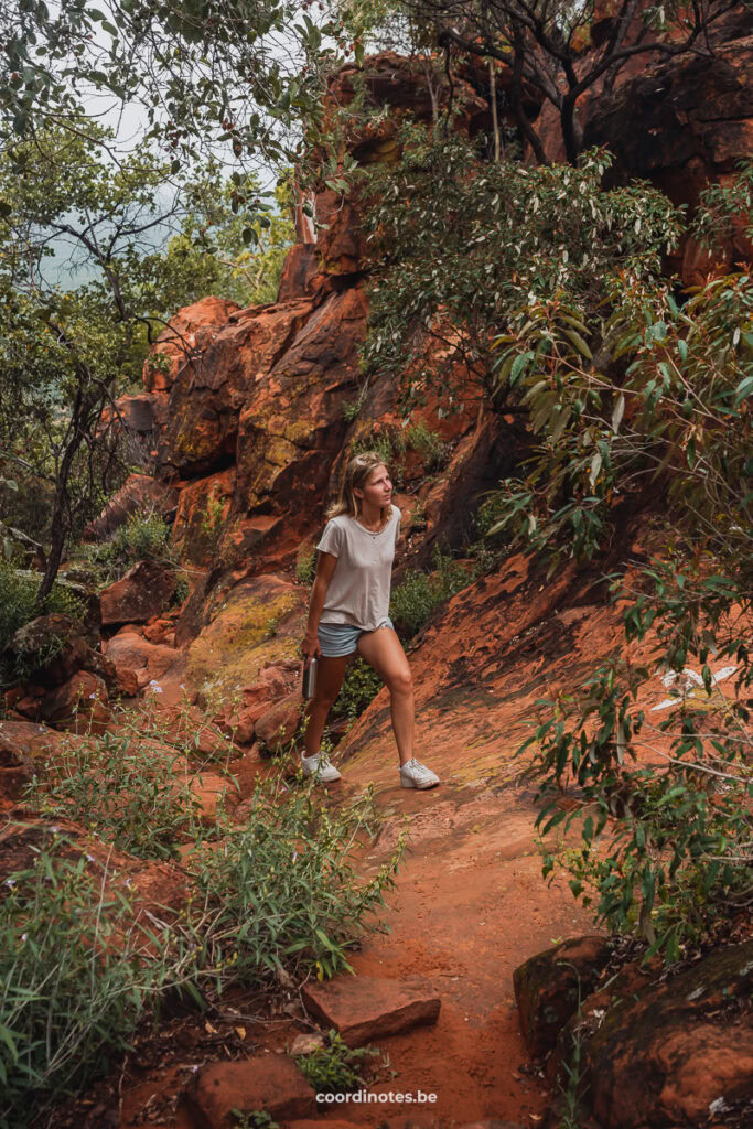 Sarah die naar boven wandelt langs een pad op een berg met een oranje ondergrond en oranje rotsen tussen de groene bosjes.