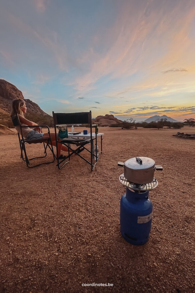 Sarah sitting in a camping chair next to another camping chair and a table. In front, there's a gas bottle with cooking pot on tob and a magical sunset over the sand plane in the backgroud.