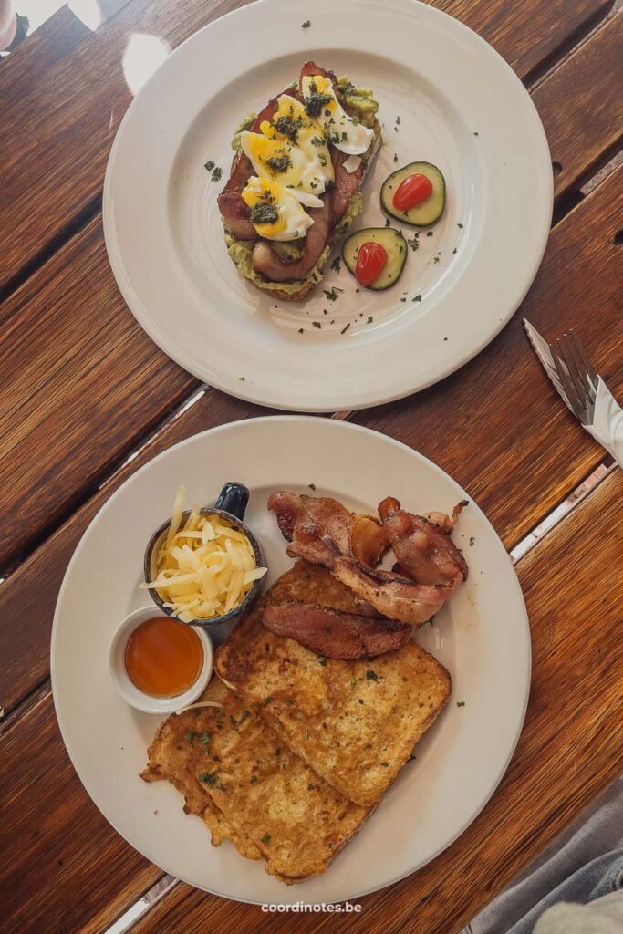 Twee bordjes met ontbijt. Op één bordje licht een toast met avocado en eitjes. Op het andere bord wentelteefjes met spek en een potje met geraspte kaas en een potje met saus.