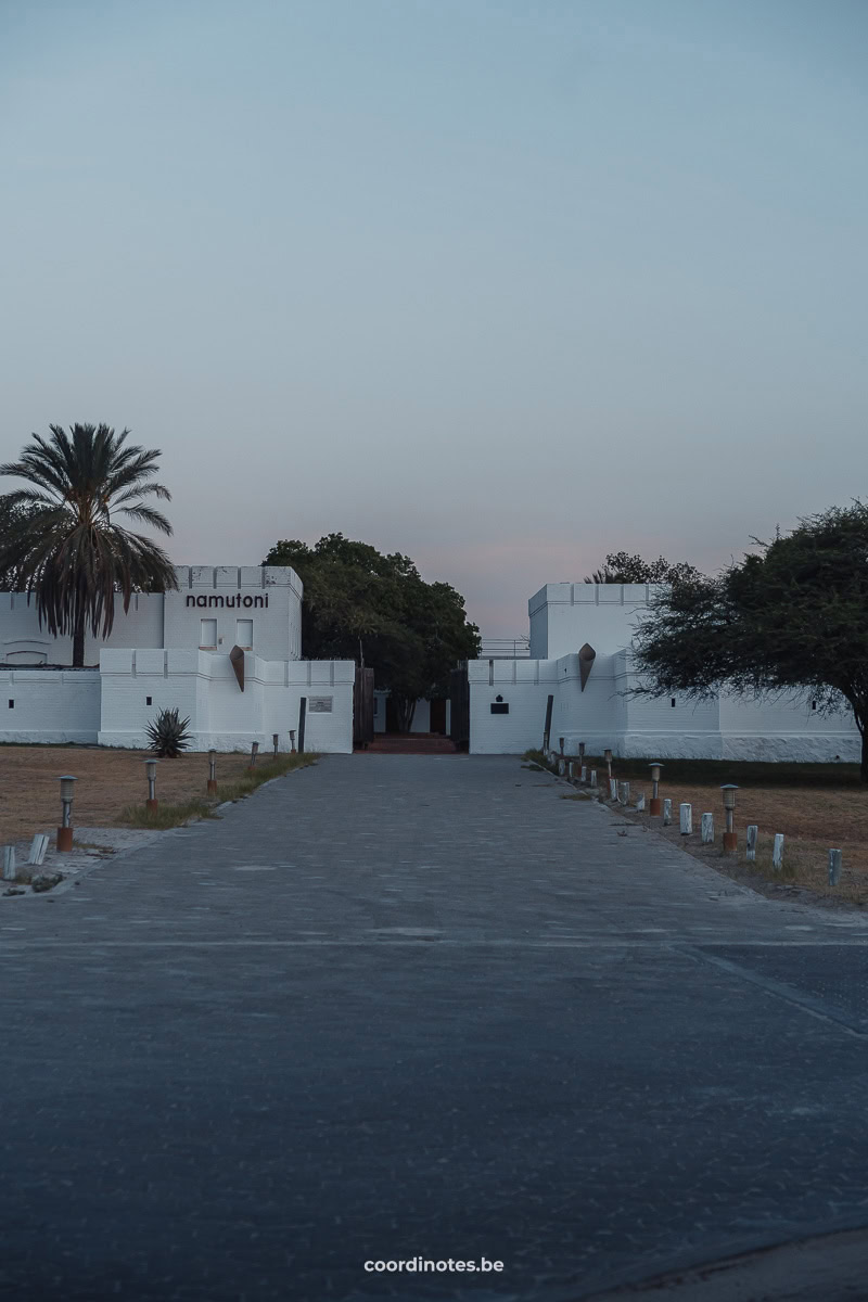 A drive way leading to a gate with white castle-like walls, with trees in the background.