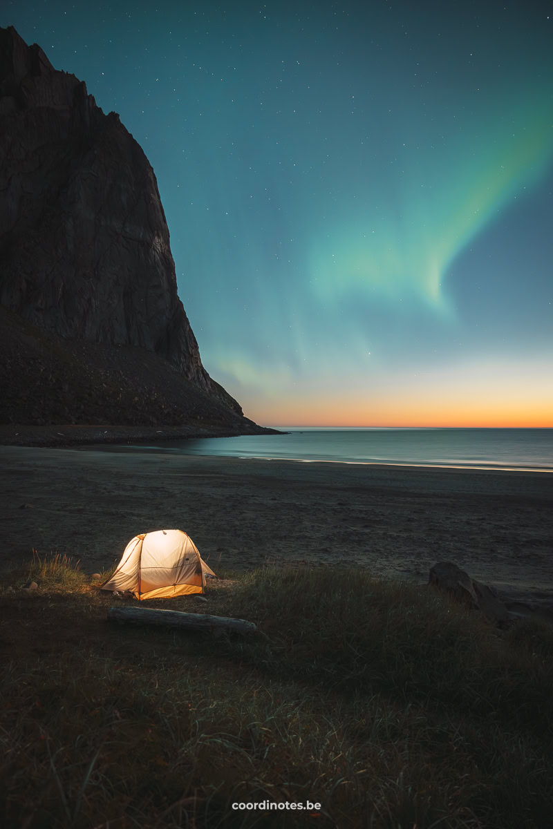 Een tentje op Kvalvika Beach waar ligt in schijnt met op de achtergrond een grote berg en het noorderlicht in de lucht tijdens het laatste licht van de zonsondergang op Kvalvika Beach.