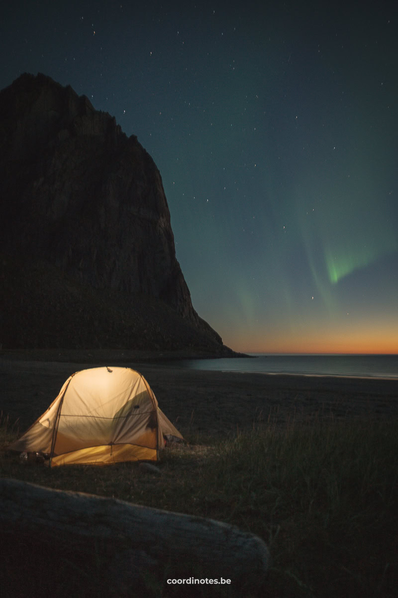 A yellow illuminated tent on the beach with a mountain in the background and the northern lights in the sky during sunset on iKvalvika Beach