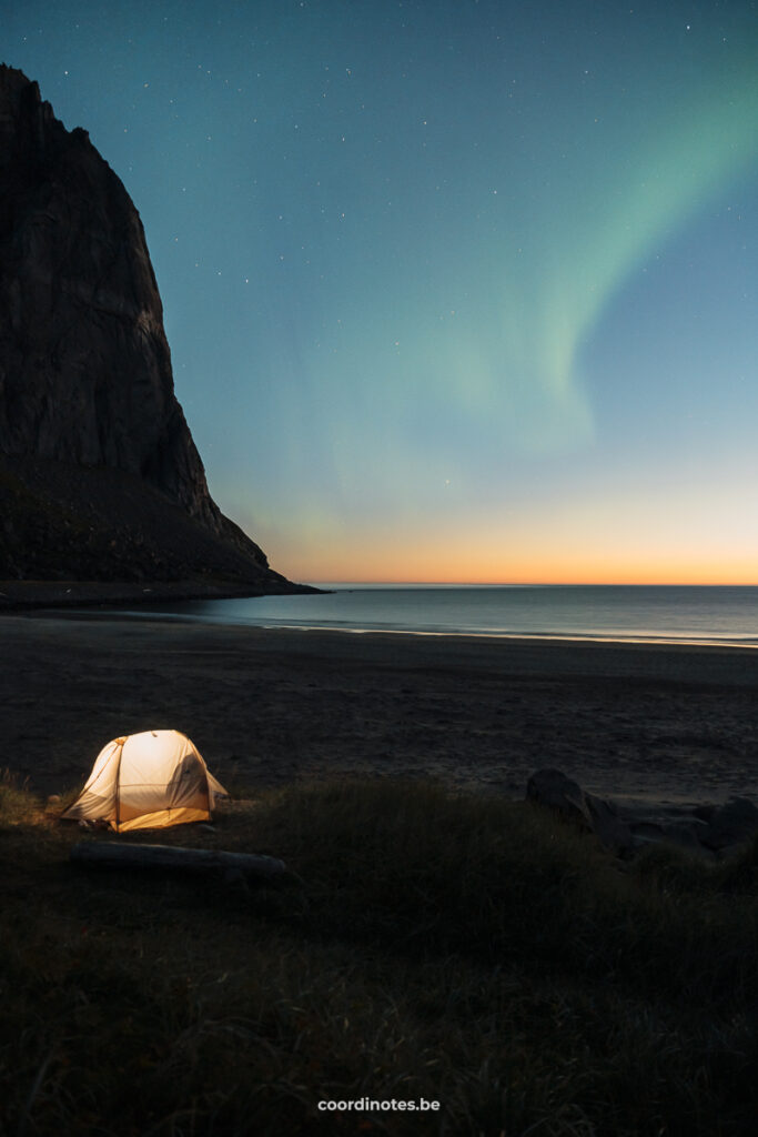 A yellow illuminated tent on the beach with a mountain in the background and the northern lights in the sky during sunset on iKvalvika Beach