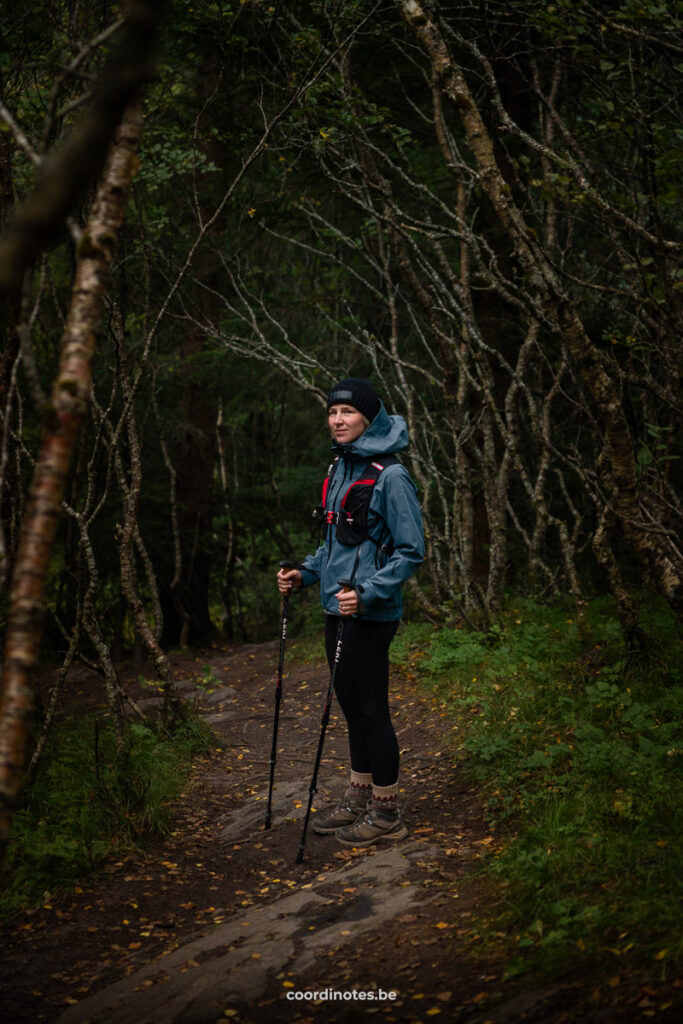 Woman in a blue hiking jacket and beanie stands on a forest trail with trekking poles.