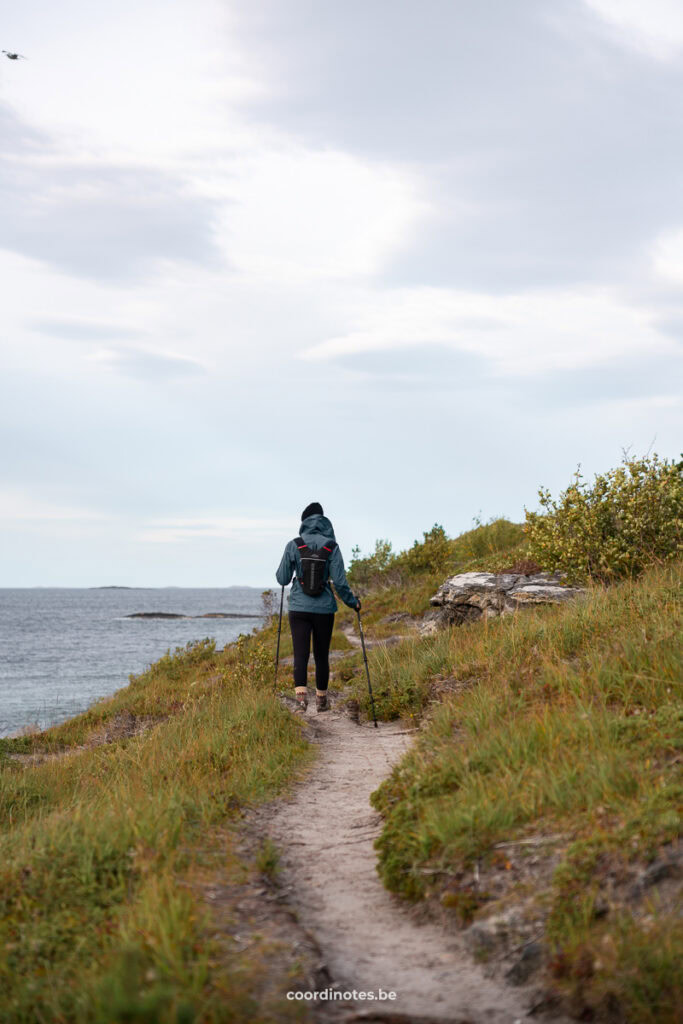 Hiker in a teal jacket with a backpack and trekking poles walking along a narrow coastal dirt trail by the sea.