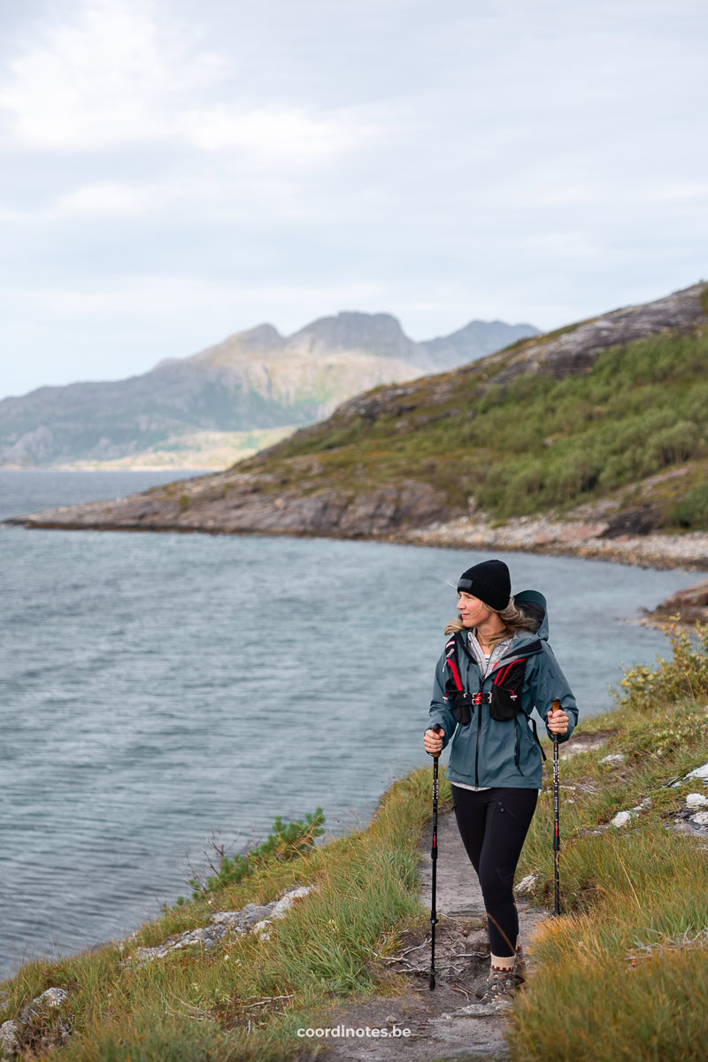 Woman hiking on a rocky coastal trail with trekking poles, wearing a teal jacket and black beanie, near the water and distant mountains.