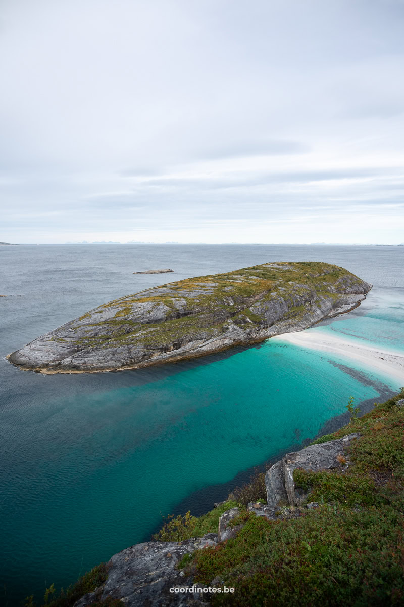 Rocky island stretching into turquoise water, mossy top, under a cloudy sky; a calm coastal scene.