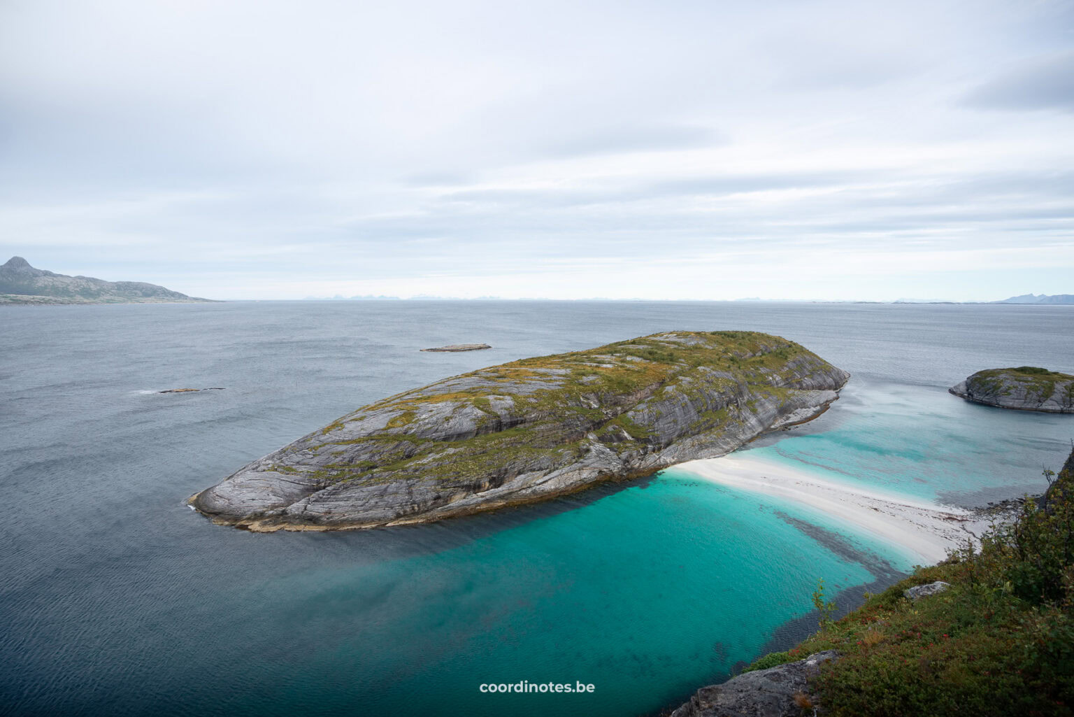 Rocky coastal island jutting into turquoise water under a pale cloudy sky, with calm sea and distant landmasses on the horizon.