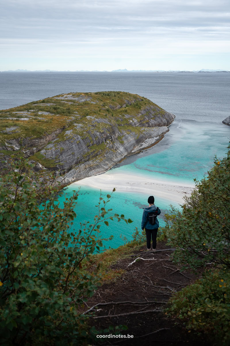Person stands on a rocky cliff overlooking an turquoise bay with white sand and distant islands under a cloudy sky.