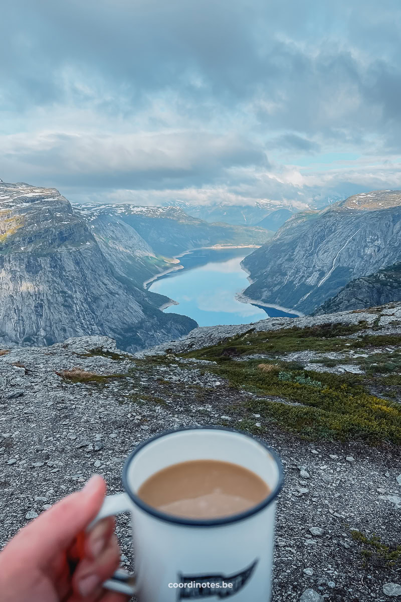 A hand holding a cup of coffee with a view over Ringedalsvatnet lake surrounded by mountains in the background at Trolltunga.