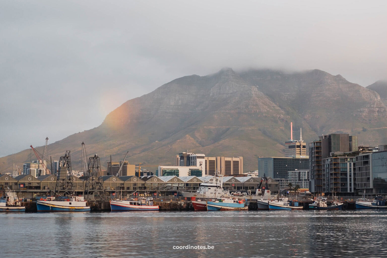 De haven bij V&A Waterfront met boten aan de kade, een berg op de achtergrond en een regenboog