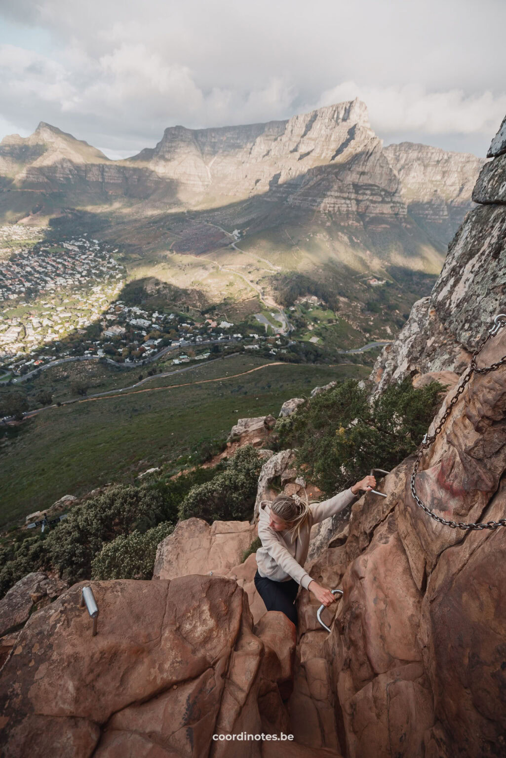 Sarah die naar boven klimt op Lions Head Mountain via metalen pinnen met Kaapstad en de Tafelberg op de achtergrond