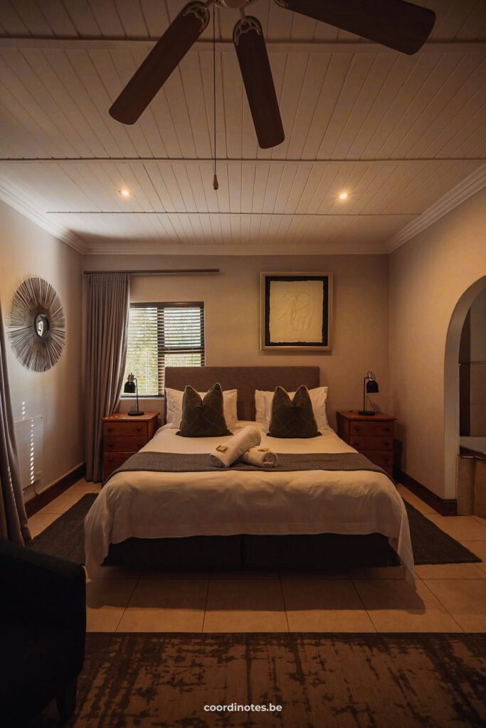 A hotelkamer in Knysna Elephant Park met een dubbel bed, een ventilator aan het plafond, een klein raam links boven het bed en decoratie aan de muur.