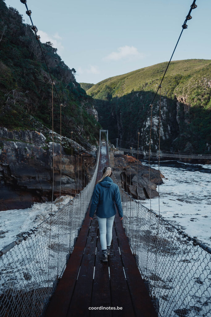 Sarah die over een hangbrug wandelt in het Tsitsikamma nationaal park over het water naar een berg in de achtergrond