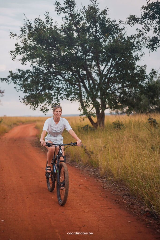 Sarah aan het fietsen over een oranje zandweg naast het hoge grass met bomen op de achtergrond in Eswatini