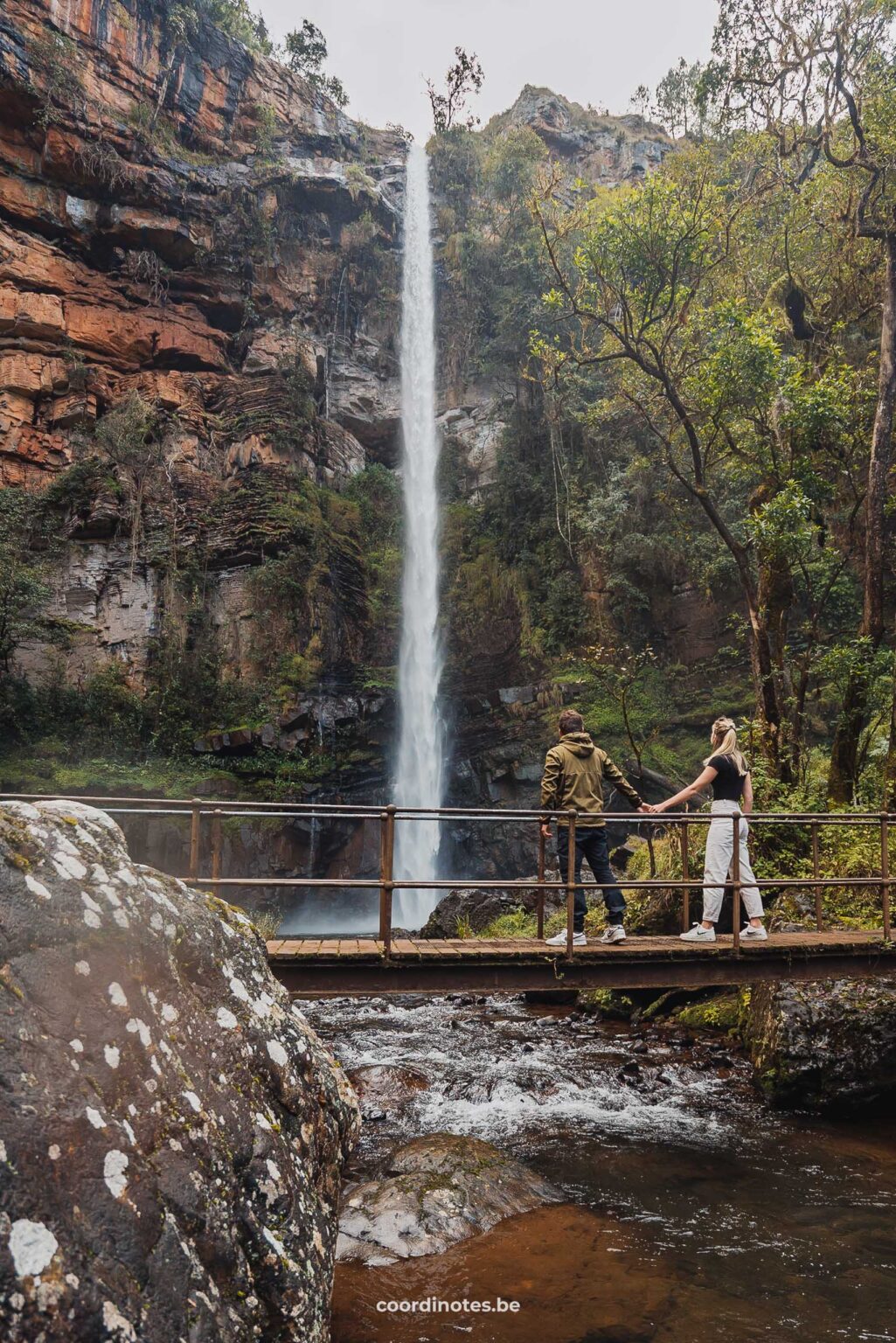 Wij die samen over een klein bruggetje wandelen voor de Lone Creek Falls waterval die van een grote oranje klif valt begroeit met mos en omgeven door bomen.