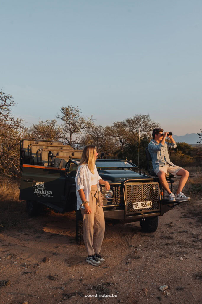 Sarah die tegen een safari jeep leunt terwijl Cédric op de spotter stoel zit terwijl hij rondkijkt met een verrekijker tijdens een Game Drive met Rukiya Safari Camp