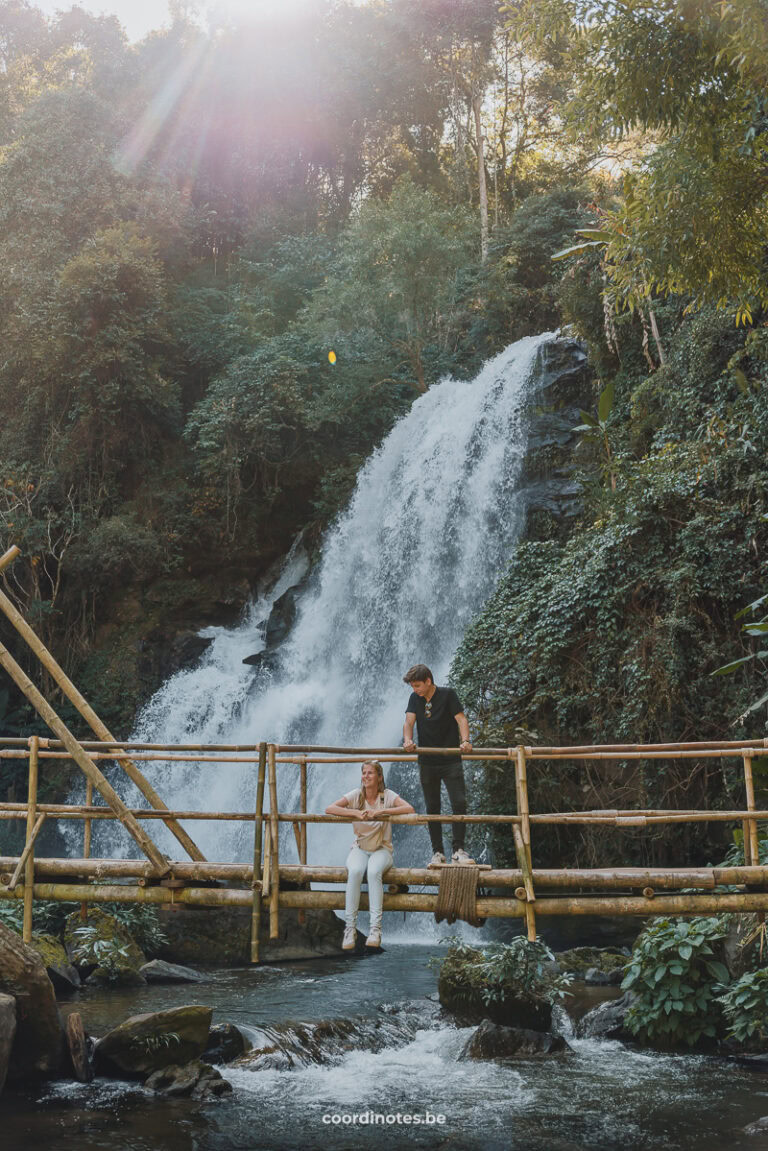 Sarah sitting down and Cédric standing up, holding the railing of the Bamboo bridge in front of a waterfall in Doi Inthanon National Park.