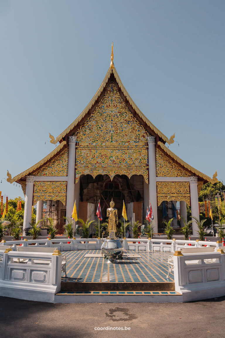 A statue of a buddha on a round blue tile floor in front of a golden triangle Thai temple.