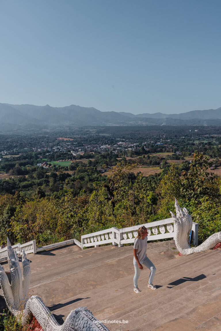 Sarah walking up a stairs with white dragons as railings, with a wide landscapes and some rolling hills in the background.