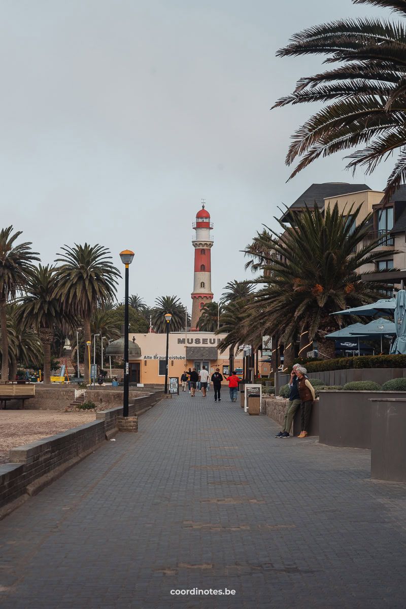 Een rood met wit gestreepte vuurtoren achter een geel gebouw op het einde van een boulevard met palmbomen in Swakopmund.