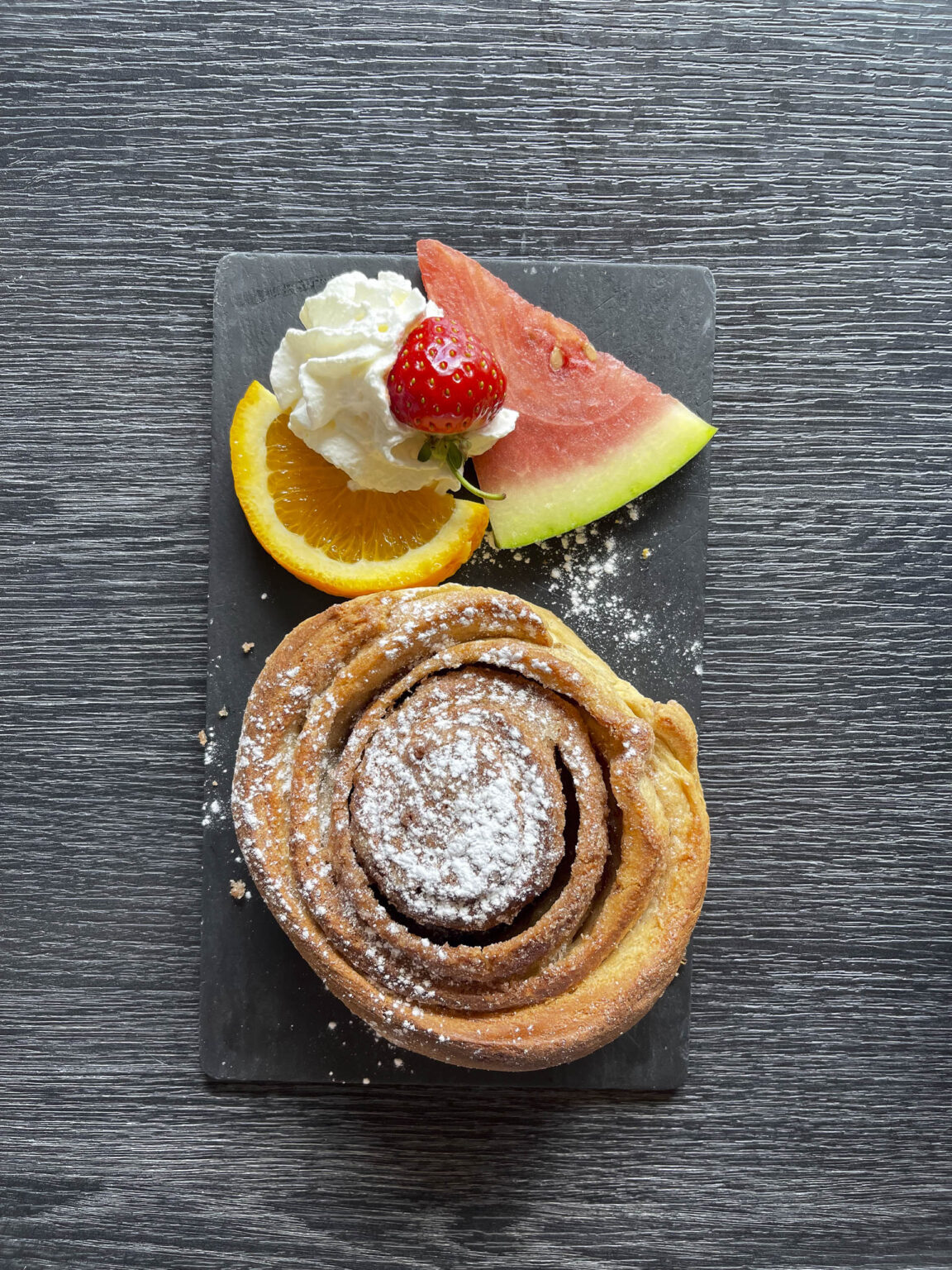 Een kaneelbol, een stukje watermeloen en een schijfje appelsien op een zwarte leisteen